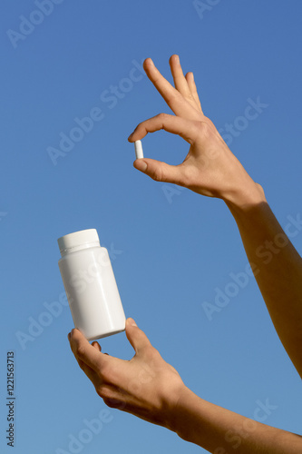 Vertical image of female hands holding white mockup jar of pills and pill capsule on blue sky background. Concept of healthcare, medicine, pharmacy, dietary supplements, disease prevention