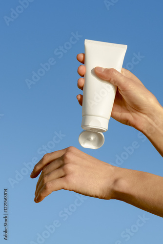 Vertical image of female hands squeezing cream from a white empty tube onto skin against blue sky background. Concept of cosmetology, skin care, protection with SPF