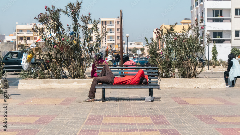 An unknown man black african man in red shirt and brown trowsers is taking a nap on a metal bench in a park in Dakar, Africa..