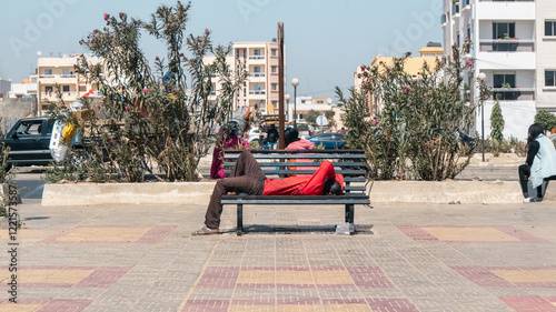 Obraz na płótnie An unknown man black african man in red shirt and brown trowsers is taking a nap on a metal bench in a park in Dakar, Africa.. w salonie