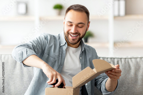 Satisfied buyer. Portrait of happy Caucasian man receiving package, unboxing parcel, taking out gaming joystick, sitting on sofa in living room. Millennial guy feeling happy with online purchase photo