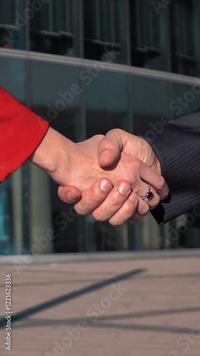Two Business partners men in black suit and women in red bright dress doing a handshake with glass business centre at the background. Slow motion close up. Vertical Shot.