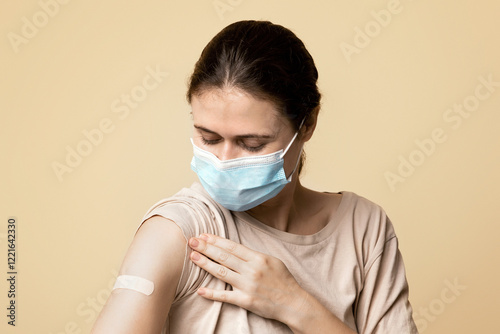 Woman in protective face mask looking at medical plaster on her shoulder, female patient got vaccinated against coronavirus at clinic or vaccination point, beige studio background, copy space photo