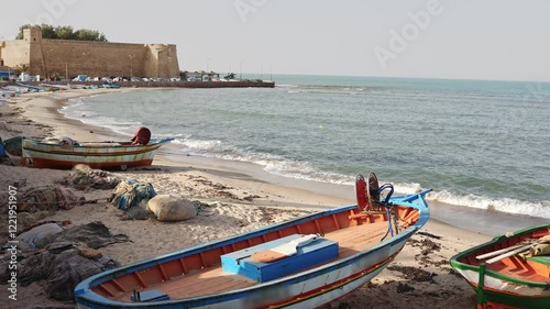 Ancient fortified citadel of Hammamet with formidable stone walls standing guard over tranquil Mediterranean seashore with old fishing boats of locals, Tunisia