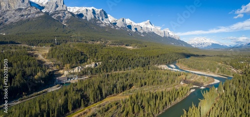Aerial view of a river winding through a valley, surrounded by lush forests and snow-capped mountains. A power plant and railway bridge are visible. , Canmore, Alberta, Canada
