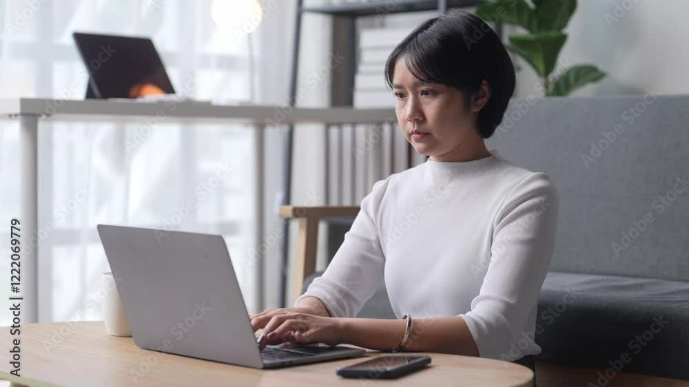 A woman is sitting at a table with a laptop and a cell phone. She is typing on the laptop and looking at the cell phone A woman is sitting at a table with a laptop and a cell phone. She is typing on the laptop and looking at the cell phone