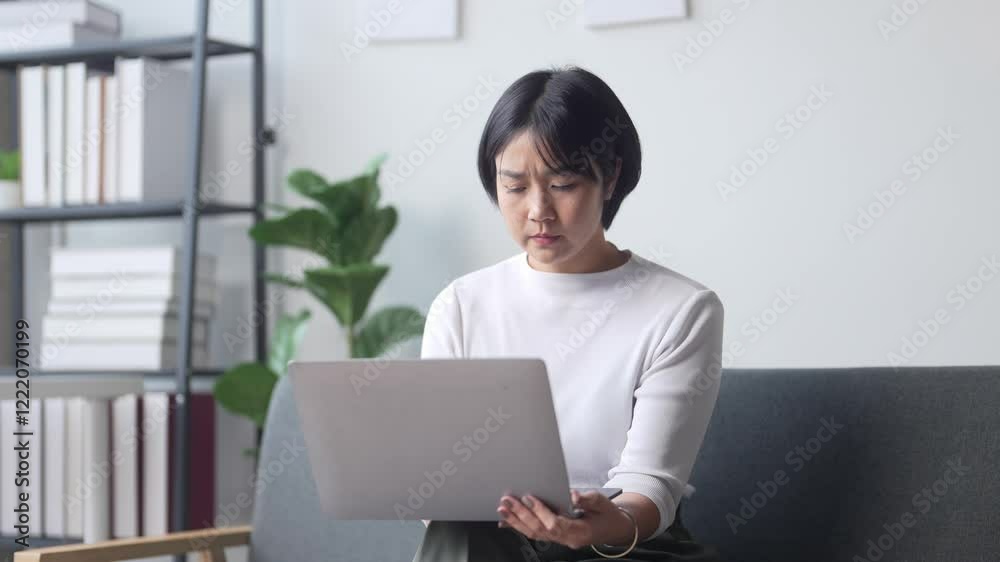 A woman is sitting on a couch and using a laptop. She is looking at the screen with a serious expression. The room has a lot of books and a potted plant A woman is sitting on a couch and using a laptop. She is looking at the screen with a serious expression. The room has a lot of books and a potted plant