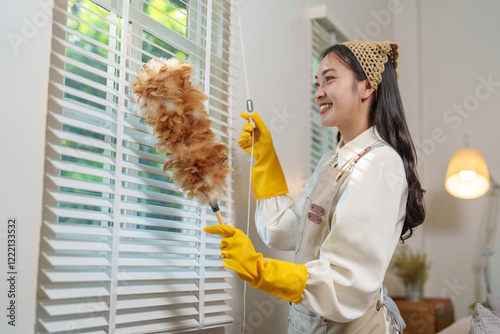 Young housekeeper with yellow gloves and an apron is smiling while cleaning venetian blinds with a feather duster in a sunlit room, performing her daily chores photo