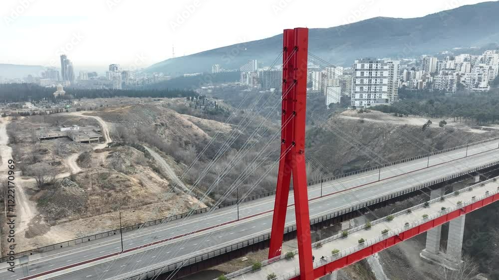 A striking red bridge spans across a wide valley, connecting urban development to distant hills. The blend of manmade infrastructure with natural beauty creates a unique visual contrast