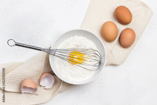 Whisk, yolk and flour in bowl. Chicken eggs on beige napkin