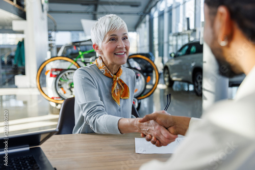 Happy senior woman shaking hands with salesperson at desk in car showroom photo