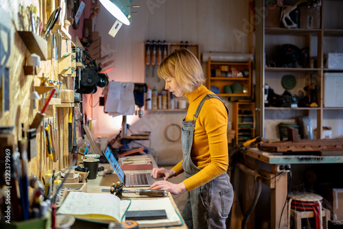 Successful female carpenter fulfill client requests on laptop in workshop. Coordinate materials, manage finances, maintaining online orders, preparing for clients or personal project for wood shop photo