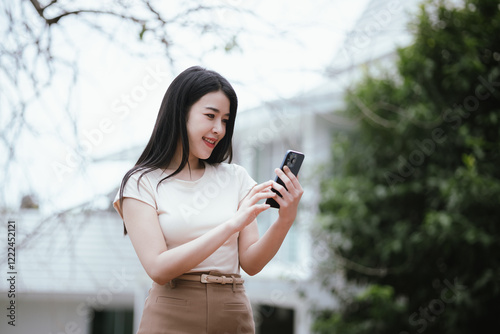 A smiling young woman is enjoying a relaxing day in a green city park, using her smartphone to work or relax. She is surrounded by nature. She works remotely while living a modern and active life. photo