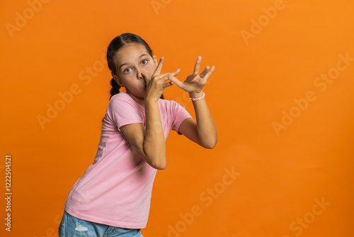 Cheerful funny bully young cute girl making faces at camera, fooling around, joking, aping with silly face, teasing. Preteen child kid 10 years old on orange background. Little children
