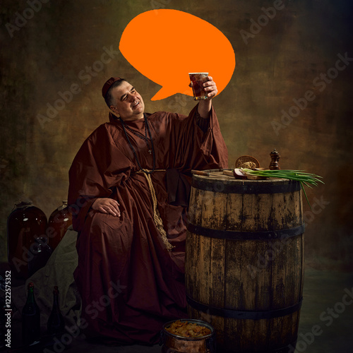 Man, medieval monk lifting glass of beer, pint of ale against dark vintage background with orange speech bubble for customizable messaging. Concept of comparison of eras, history, brewery, traditions photo