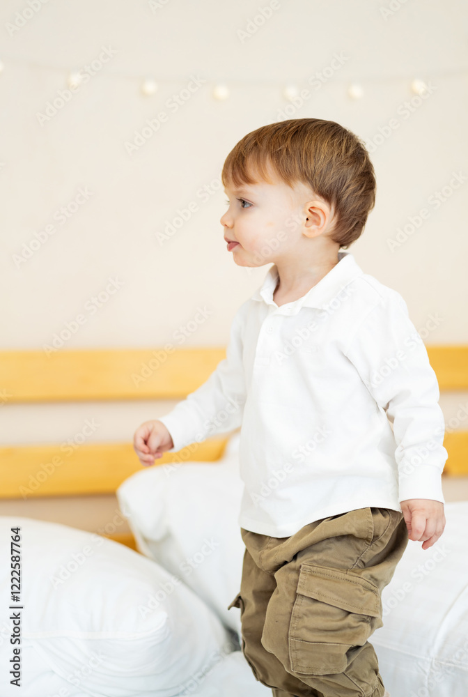 Naklejka premium Toddler playing and exploring indoors. A young child joyfully stands on a bed, looking around with curiosity in a bright room filled with soft colors.