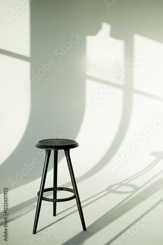 A black tall stool on three legs in a white studio bathed in light. Artist's studio, minimalism