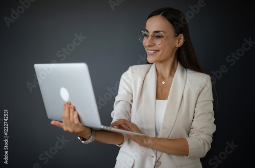 A confident businesswoman in a white suit holds a laptop while smiling. She wears glasses and is in a modern office setting, conveying professionalism, productivity, and success. photo