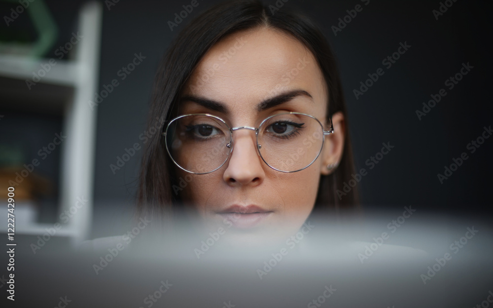 The image shows close-up of a focused woman wearing glasses, working on laptop. She appears deeply concentrated, with professional demeanor, in modern office space with bookshelves in the background The image shows close-up of a focused woman wearing glasses, working on laptop. She appears deeply concentrated, with professional demeanor, in modern office space with bookshelves in the background