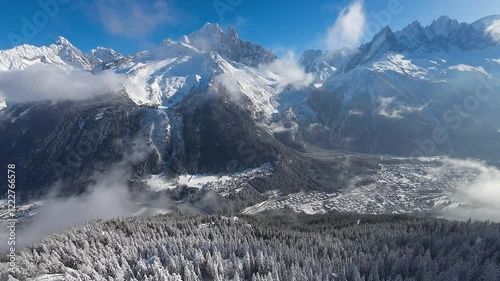 Aerial drone view of Chamonix’s ski slopes, surrounded by the towering white peaks of the Alps, offering a stunning winter wonderland with white forests and the town under a bright midday 