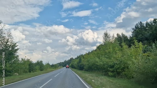 A first-person view of the car behind the wheel. A road with trees and woods. Blue sky on a sunny afternoon.