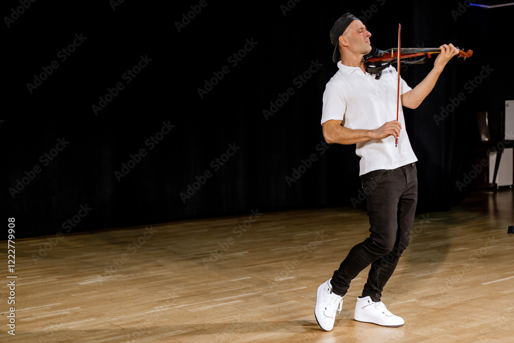40 years old man in white shirt and black cap standing on the black background having joy while playing violin, musical instrument in scene. Male violinist, copy space 40 years old man in white shirt and black cap standing on the black background having joy while playing violin, musical instrument in scene. Male violinist, copy space