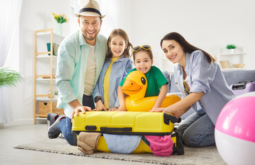 Happy family packing bright yellow suitcase in living room for upcoming fun trip. Parents and children smiling, organizing clothes and toys, preparing for journey. Traveling and vacation concept.