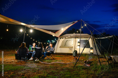 Outdoor camping tent in the forest park, party dinner with friends under tarp or flysheet and warm lighting at night near natural under a twilight sky. Overview of travel camping with friends. photo