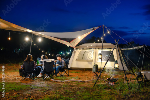 Outdoor camping tent in the forest park, party dinner with friends under tarp or flysheet and warm lighting at night near natural under a twilight sky. Overview of travel camping with friends.
