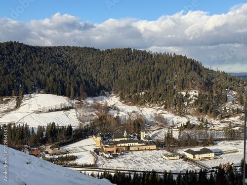 A beautiful winter landscape features snow-covered hills alongside towering pine trees and a Christian monastery