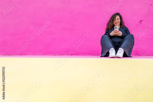 Pregnant woman smiling, browsing smartphone while resting on ledge near pastel colored wall background