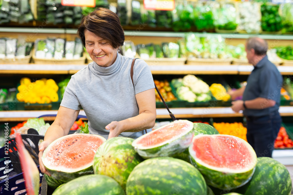 Obraz premium Elderly woman chooses watermelon in fruit and vegetable section