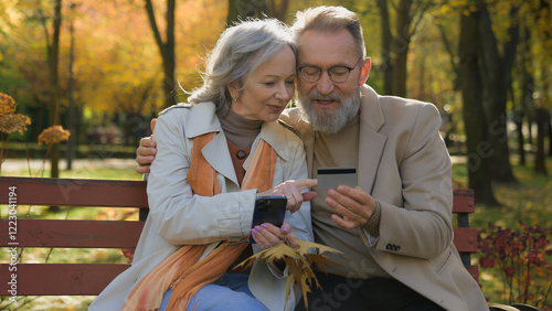Married Caucasian couple happy family middle-aged mature woman man remote paying online order with mobile phone credit card retired husband wife using smartphone bank app shopping in autumn park city