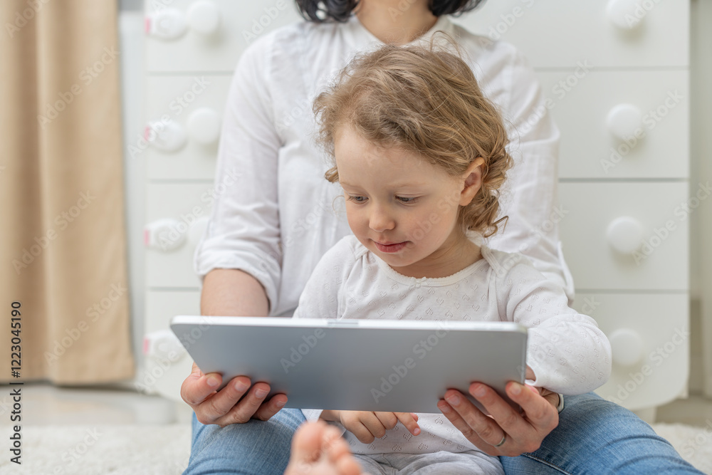 A young girl child with curly hair is sitting on her parent's lap, looking attentively at a tablet screen. The scene depicts a tender moment of learning and bonding in a cozy indoor setting.