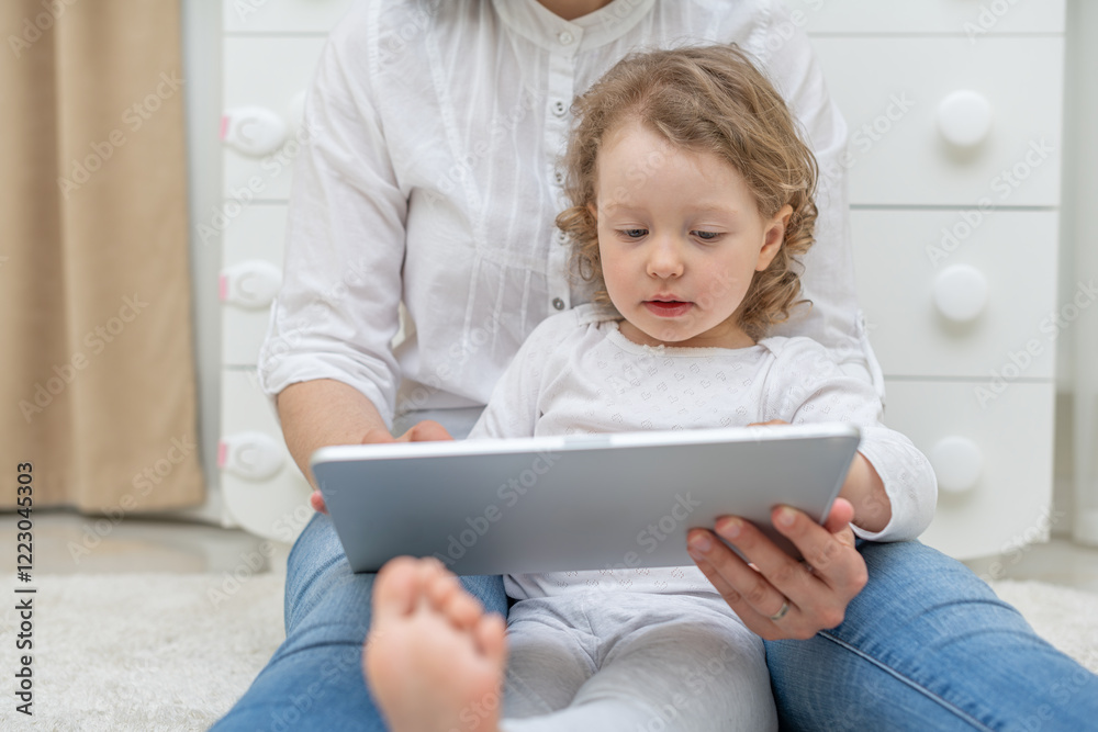 A young girl child with curly hair is sitting on her parent's lap, looking attentively at a tablet screen. The scene depicts a tender moment of learning and bonding in a cozy indoor setting.