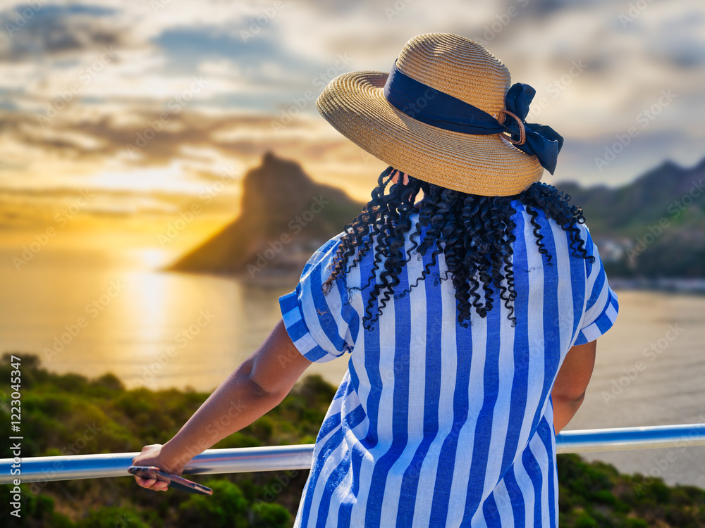 African woman in a blue and white striped dress standing and enjoying the sea view at sunset in Hout Bay, Cape Town, South Africa