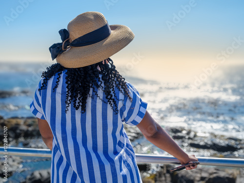 African woman in a blue and white striped dress standing and enjoying the sea view at sunset in Green Point, Cape Town, South Africa