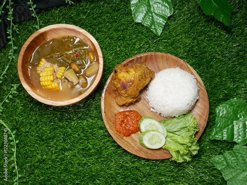 Complete traditional Indonesian food featuring fried chicken, white rice, red sambal, cucumber slices, and fresh lettuce. Sayur asam in a wooden bowl with corn, chayote, long beans, and melinjo seeds. photo