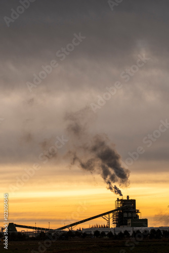 An industrial factory belches thick smoke into the sky at sunset, illustrating concerns about pollution and environmental impact in an urban landscape.