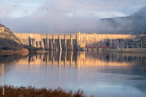 A grand dam stands tall against a misty backdrop, with water flowing beneath it, reflecting the structure and surrounding nature, depicting human achievement in engineering. photo