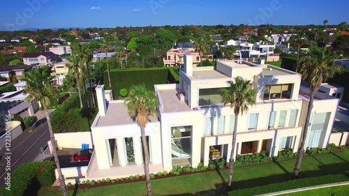 Aerial view of a large villa in Melbourne surrounded by palm trees, with the city skyline in the background. Bright and sunny atmosphere showcasing luxury living. Drone move left and up.
