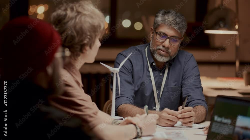 Mature male engineer talking about renewable energy strategies with team of colleagues, sitting at table with wind turbine model on it during meeting focused on sustainability and innovation