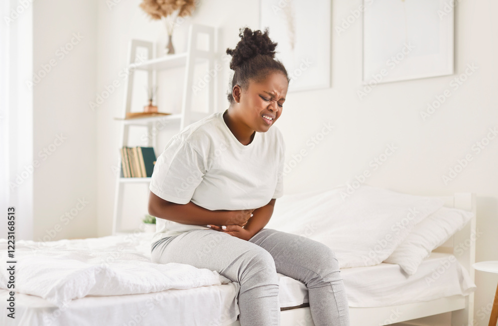 Sick african american woman sitting on bed putting hands on belly, suffering from strong bad stomach ache, unhappy girl having painful feeling in abdomen, pancreatitis gastritis symptom, appendicitis Sick african american woman sitting on bed putting hands on belly, suffering from strong bad stomach ache, unhappy girl having painful feeling in abdomen, pancreatitis gastritis symptom, appendicitis