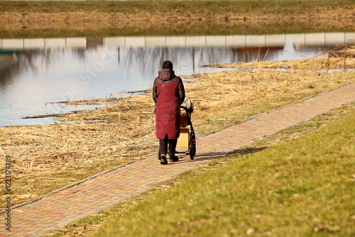 A young mother walks in the park with a baby carriage on a sunny day. Baby's stay in the fresh air. Raising the younger generation. Guardianship of human offspring.