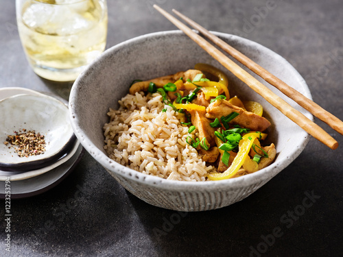 Stir-fried chicken strips mixed with fresh vegetables sit atop a bed of brown rice. A glass of chilled drink and small plates with spices complete the table setting photo