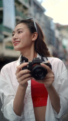 Vertical portrait of cheerful young traveler woman using camera while standing side of bangkok street in Thailand, female enjoy exploring and capturing memories on her adventure lifestyle holiday.
 photo