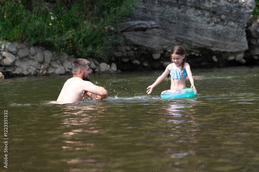 Fototapeta premium A man and young girl are playing in a calm river. The girl floats on a small inner tube. The rocky riverbank is visible in the background. It's a sunny day, and the water appears cool and inviting.