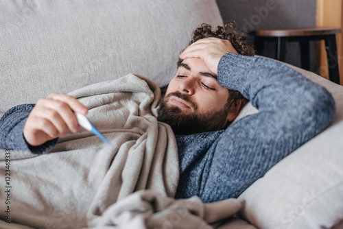Man with fever checking temperature while lying sick on a couch. cold, flu, virus season