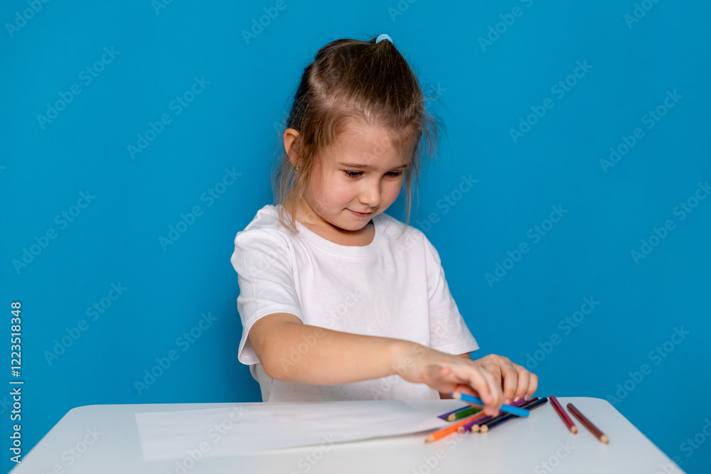 Little girl in casual white t-shirt sitting at a table and drawing on white paper with colored pencils on a blue background. Concept of children's creativity. Mock up. High quality photo Little girl in casual white t-shirt sitting at a table and drawing on white paper with colored pencils on a blue background. Concept of children's creativity. Mock up. High quality photo
