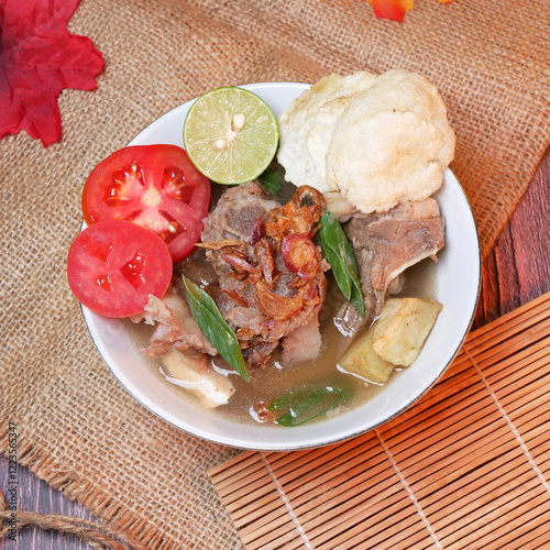 Top view of a bowl of Beef ribs soup or sop Iga with beef bone, potato, fresh tomato, lime slices, and fried shallot. Served on bamboo mats, burlap, and leaves